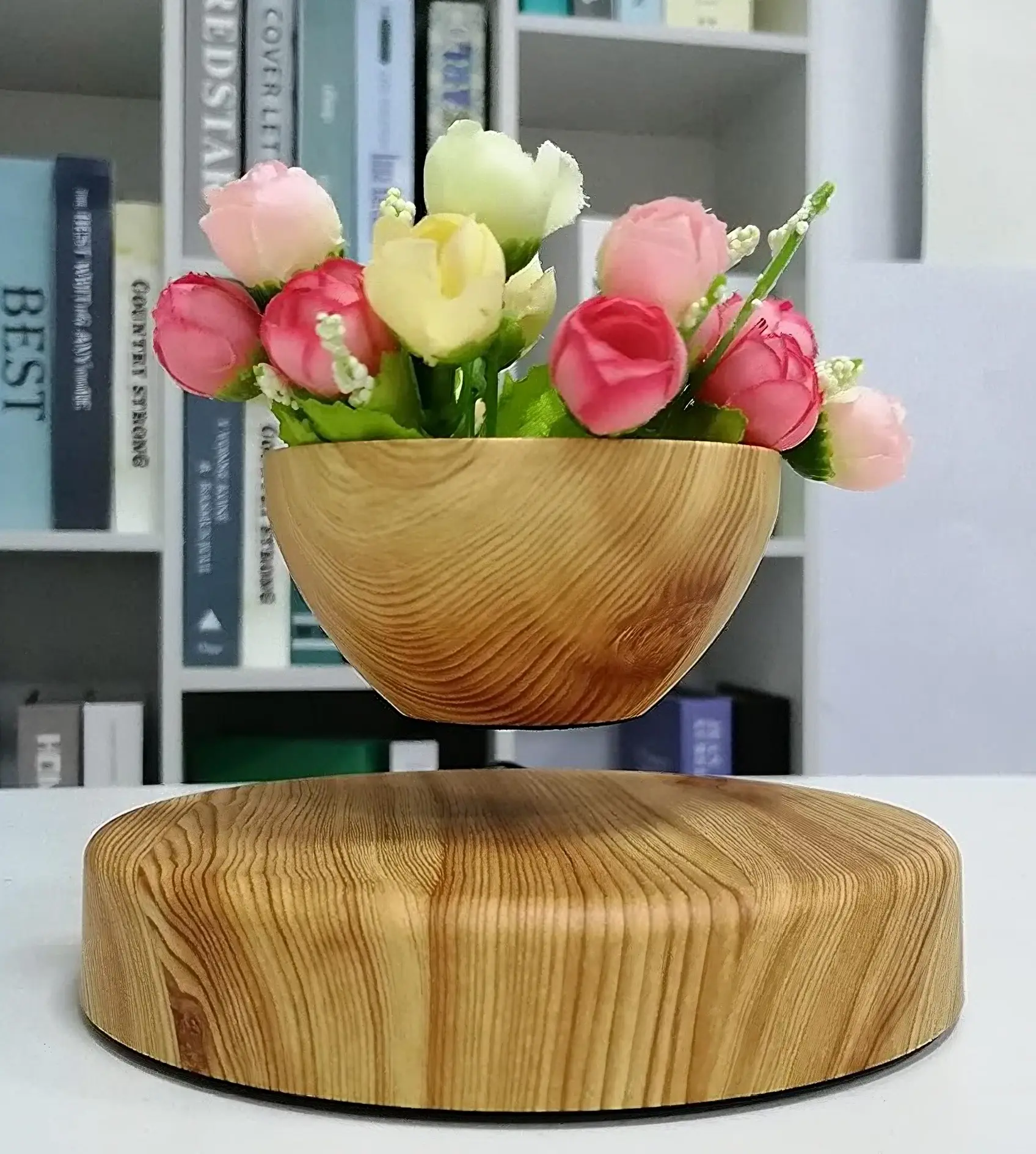 A Floating Wooden Plant Pot, accompanied by pink and white flowers, hovers gracefully above its wooden base with books in the background.