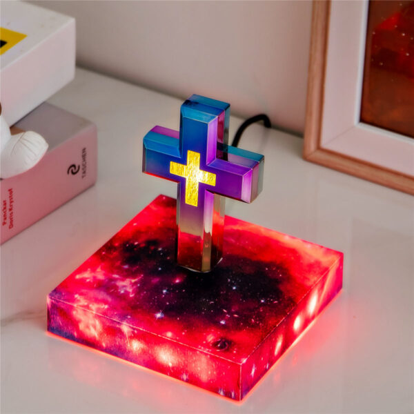 1-levitating cross nightlight with starry base The Floating Cross Nightlight, a vibrant and illuminated Christian cross, stands on a square base featuring a cosmic design. It is placed on a white surface alongside books and a framed picture.