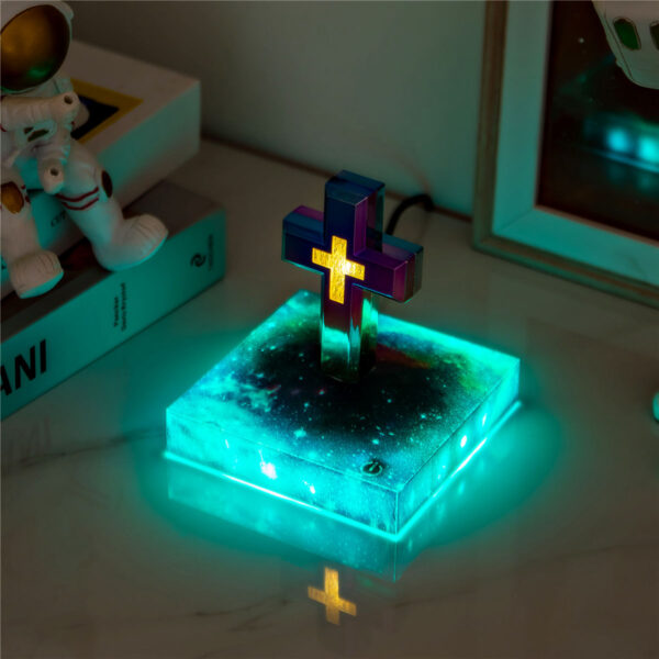 3-levitating cross nightlight with starry base The Floating Cross Nightlight, inspired by the Christian cross, is crafted from transparent material featuring a purple and yellow gradient. It stands on a book and emits turquoise light across the table.