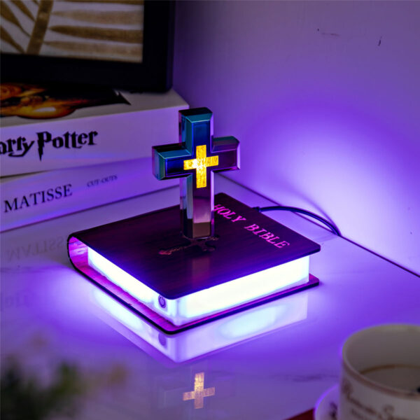 5-levitating cross nightlight with starry base A Floating Cross Nightlight on a desk emits purple light, illuminating a nearby Holy Bible, a Harry Potter book, and a cup.