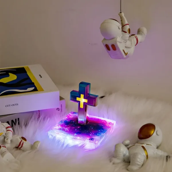 Christmas gift Personalized gifts home decoration Magnetic levitation cross floating crass table lamp RGB color 4 Decorative setup featuring astronaut figurines, a glowing cross on a galaxy-patterned base, and a book on a soft, white surface. An astronaut hangs above the scene.
