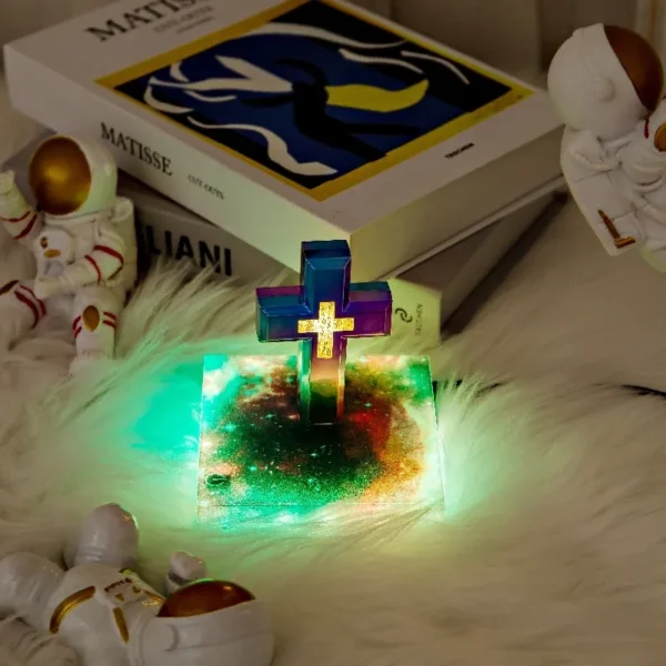 Christmas gift Personalized gifts home decoration Magnetic levitation cross floating crass table lamp RGB color 5 A glowing cross with a colorful light base is surrounded by small astronaut statues. Two books, including one by Matisse, are in the background on a fuzzy surface.