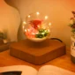 A levitating glass orb with flowers and small lights inside, sits on a wooden base on a table beside an open book.