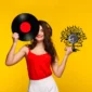 Woman in a red top holds a vinyl record and a clock with a decorative design against a yellow background.