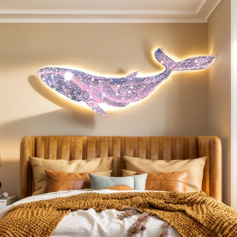 A wall-mounted, illuminated art piece of a whale with a galaxy pattern hangs above a bed with a cushioned headboard, pillows, and a knitted throw blanket.