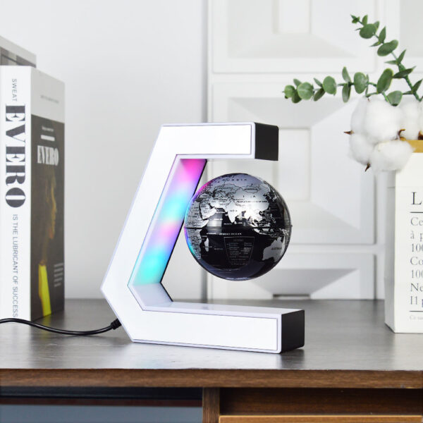 Plastic World Geography Map Rotating Globe Magnetic Levitating Floating Globe for Kids Education Home Office Desktop Decoration 4 A black and silver globe levitates inside a C-shaped magnetic stand with LED lights, placed on a wooden desk near books and a vase with green and white foliage.